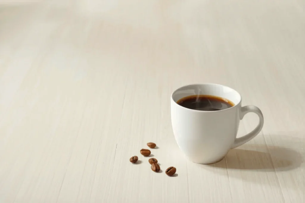 Xícara branca de café preto sobre mesa clara, com vapor leve subindo e alguns grãos de café ao lado